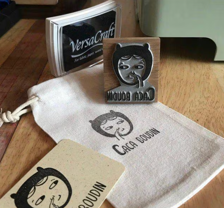 Custom rubber stamp with illustrated logo branding stamped onto a cotton drawstring bag and business card using black VersaCraft ink pad.