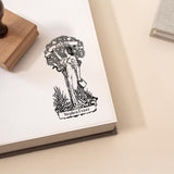 An ex libris rubber stamp featuring a beautifully detailed vintage-style illustration of a woman reaching toward a floral wreath with an Ex Libris banner being used to stamp on a white paper in a book.