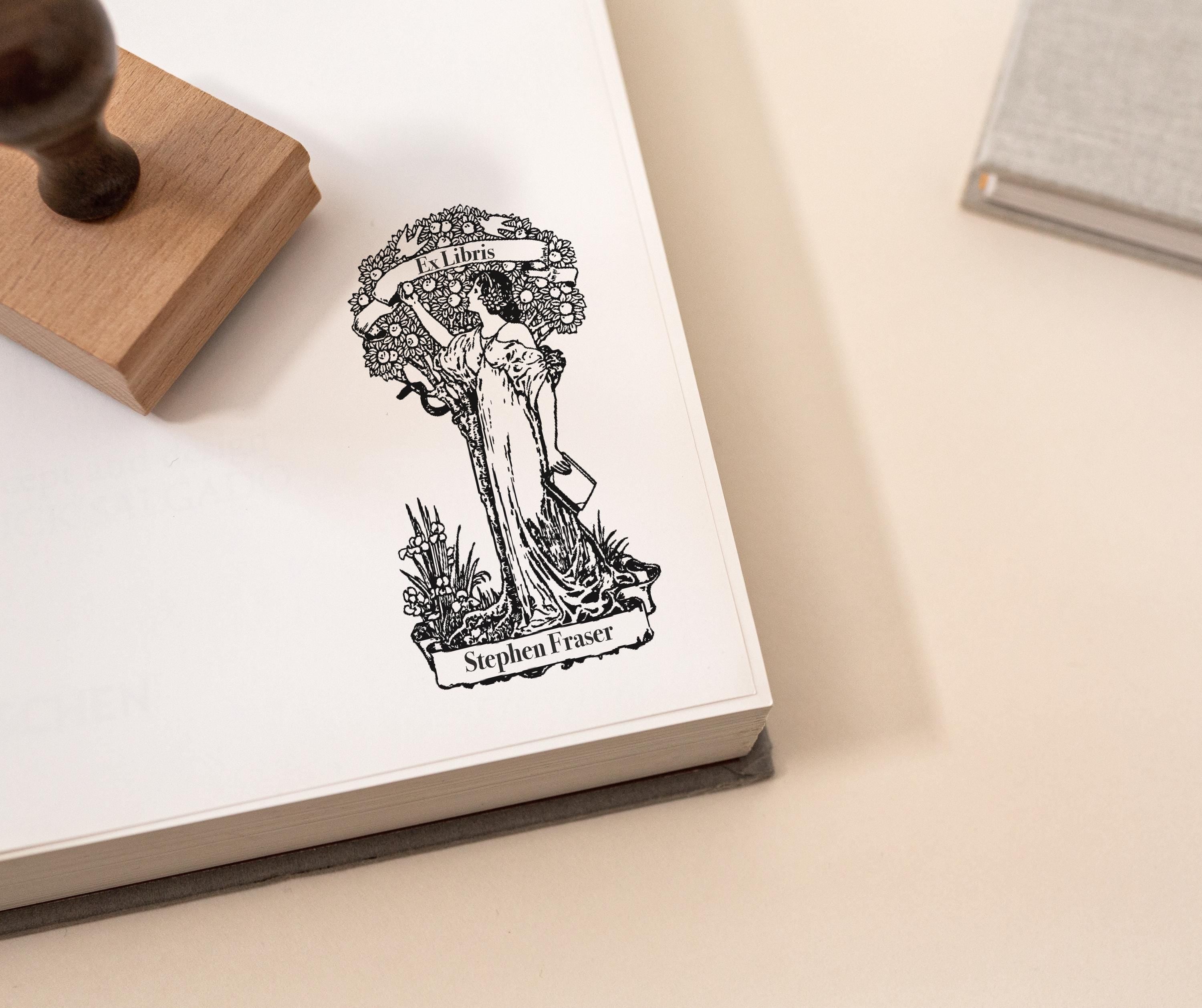 An ex libris rubber stamp featuring a beautifully detailed vintage-style illustration of a woman reaching toward a floral wreath with an Ex Libris banner being used to stamp on a white paper in a book.