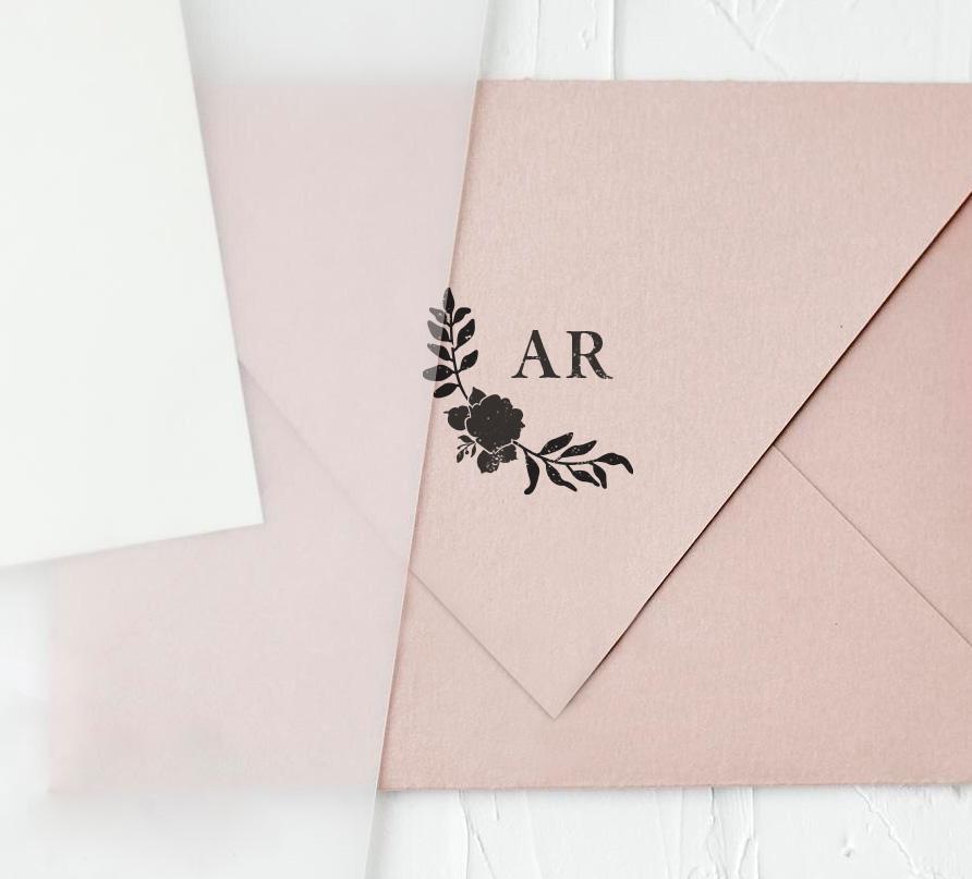 A custom rubber stamp with initials 'AR' and a floral design, pressed onto a pink envelope, creating a black monogram stamp impression.