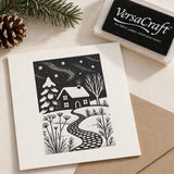 A square cream card with a black-and-white winter house illustration, shown with a VersaCraft ink pad and kraft paper envelope on a light neutral background. Pine branches and a pinecone are styled at the top left.