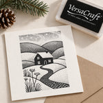A Christmas card featuring a black and white block print illustration of a cottage in a rolling landscape, styled on a beige surface with a VersaCraft ink pad and a pine cone.
