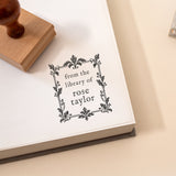 A close-up of a white book page stamped with the phrase 'from the library of rose taylor' in a decorative black frame, with a wooden rubber stamp partially visible in the upper left on a warm beige surface.