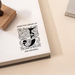 A custom library stamp with a floral monogram design being used to stamp a book cover, leaving an imprint of the design along with the text 'FROM THE LIBRARY OF Jon Deverill'.