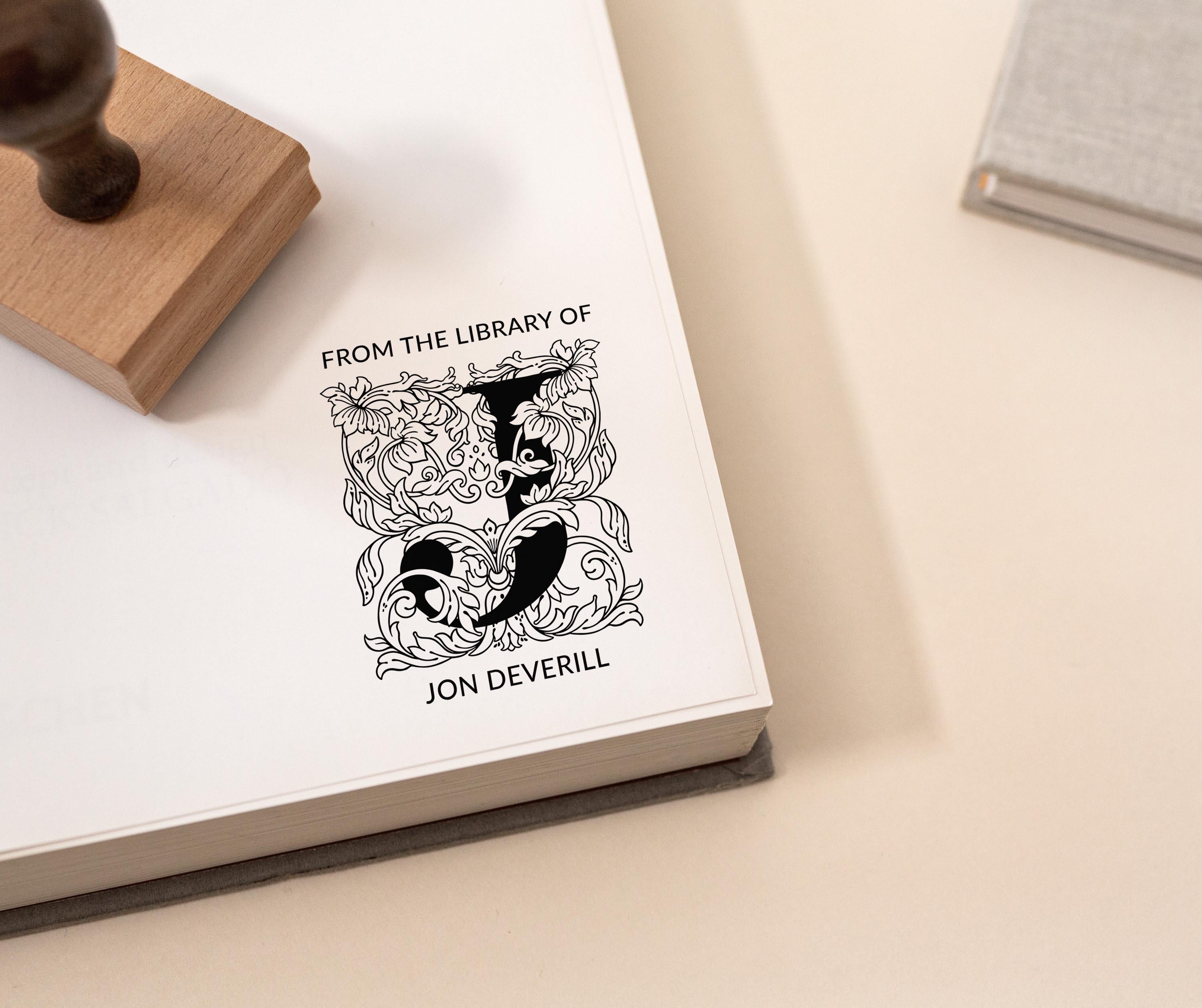 A custom library stamp with a floral monogram design being used to stamp a book cover, leaving an imprint of the design along with the text 'FROM THE LIBRARY OF Jon Deverill'.