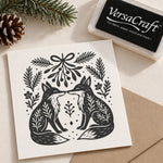 A rubber stamp and ink pad set displayed on a neutral background with a kraft paper envelope and pine decoration. The stamp features a black ink design of two foxes facing each other, surrounded by festive evergreen branches and a bow.