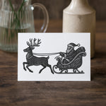 Greeting card with black and white illustration of Santa Claus in a sleigh pulled by reindeer on a wooden surface.