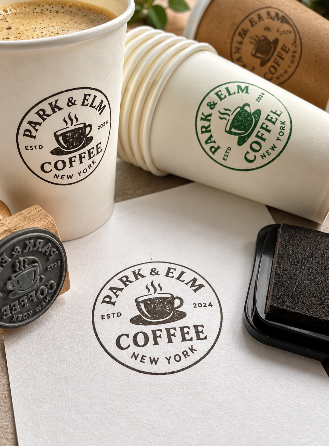 A branded coffee cup stamp set from Park & Elm Coffee, including a wooden-handled rubber stamp, a black StazOn ink pad, stacked white paper cups with green and dark gray printed logos, and a paper cup filled with coffee on a tabletop.