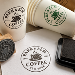 A branded coffee cup stamp set from Park & Elm Coffee, including a wooden-handled rubber stamp, a black StazOn ink pad, stacked white paper cups with green and dark gray printed logos, and a paper cup filled with coffee on a tabletop.