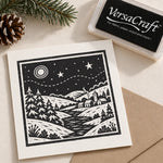 A Christmas card featuring a black and white winter landscape block print design, shown next to its black VersaCraft ink pad packaging on a beige surface with a pinecone and evergreen branch as props.