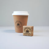 A custom coffee cup rubber stamp with an ink pad, showing a imprint of a coffee shop logo on a disposable coffee cup with a lid.