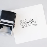 Black rubber self-inking stamp with a signature on a white card on a light gray background