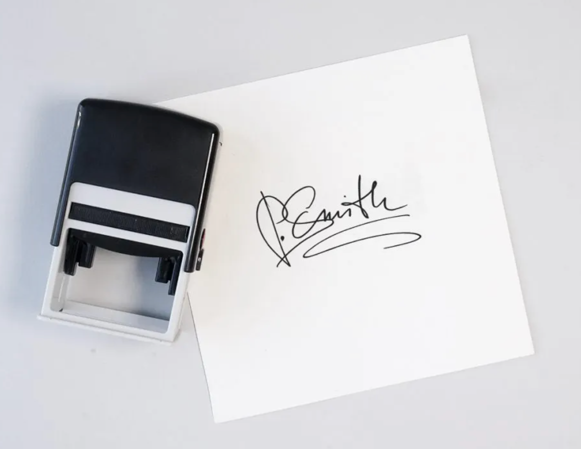 Black rubber self-inking stamp with a signature on a white card on a light gray background