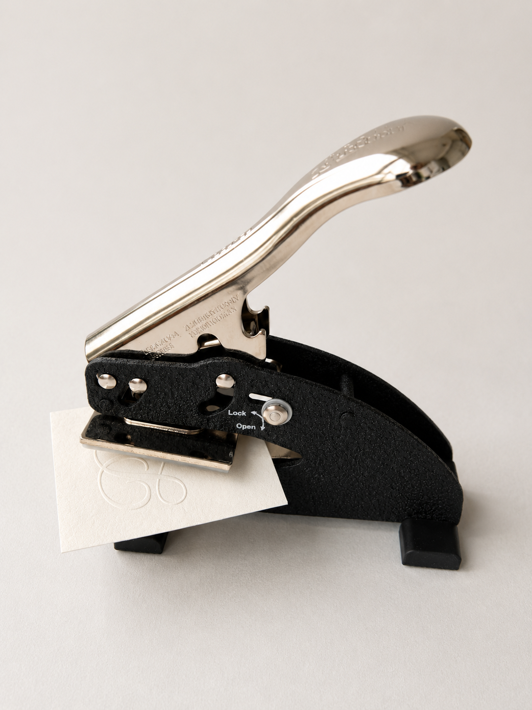 A black and silver paper embossing tool with a metallic handle, shown with a small white sheet of paper inserted with an embossed logo, on a light gray background.