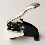 A black and silver paper embossing tool with a metallic handle, shown with a small white sheet of paper inserted with an embossed logo, on a light gray background.