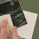 A large rubber stamp featuring a winter scene of the countryside stamping on a white christmas card