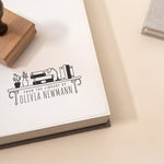 Custom book stamp with a bookshelf illustration and the wording 'from the library of' stamped onto a book and featuring a wooden rubber stamp