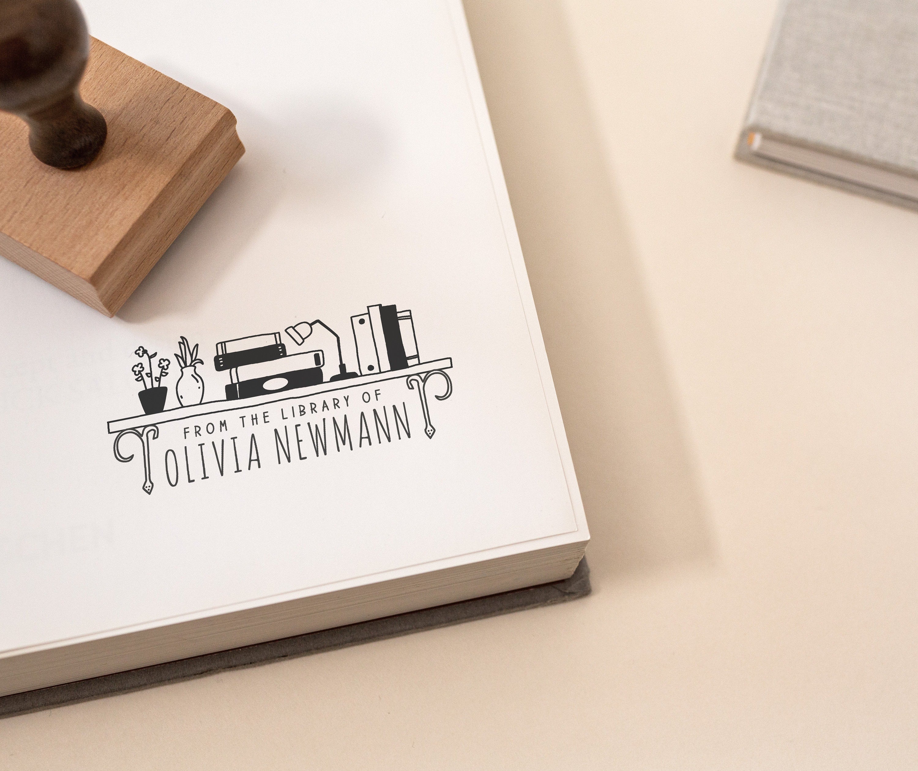 Custom book stamp with a bookshelf illustration and the wording 'from the library of' stamped onto a book and featuring a wooden rubber stamp