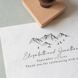 A custom wedding stamp with a mountain design being used on a piece of paper to create an impression that includes personalized text.
