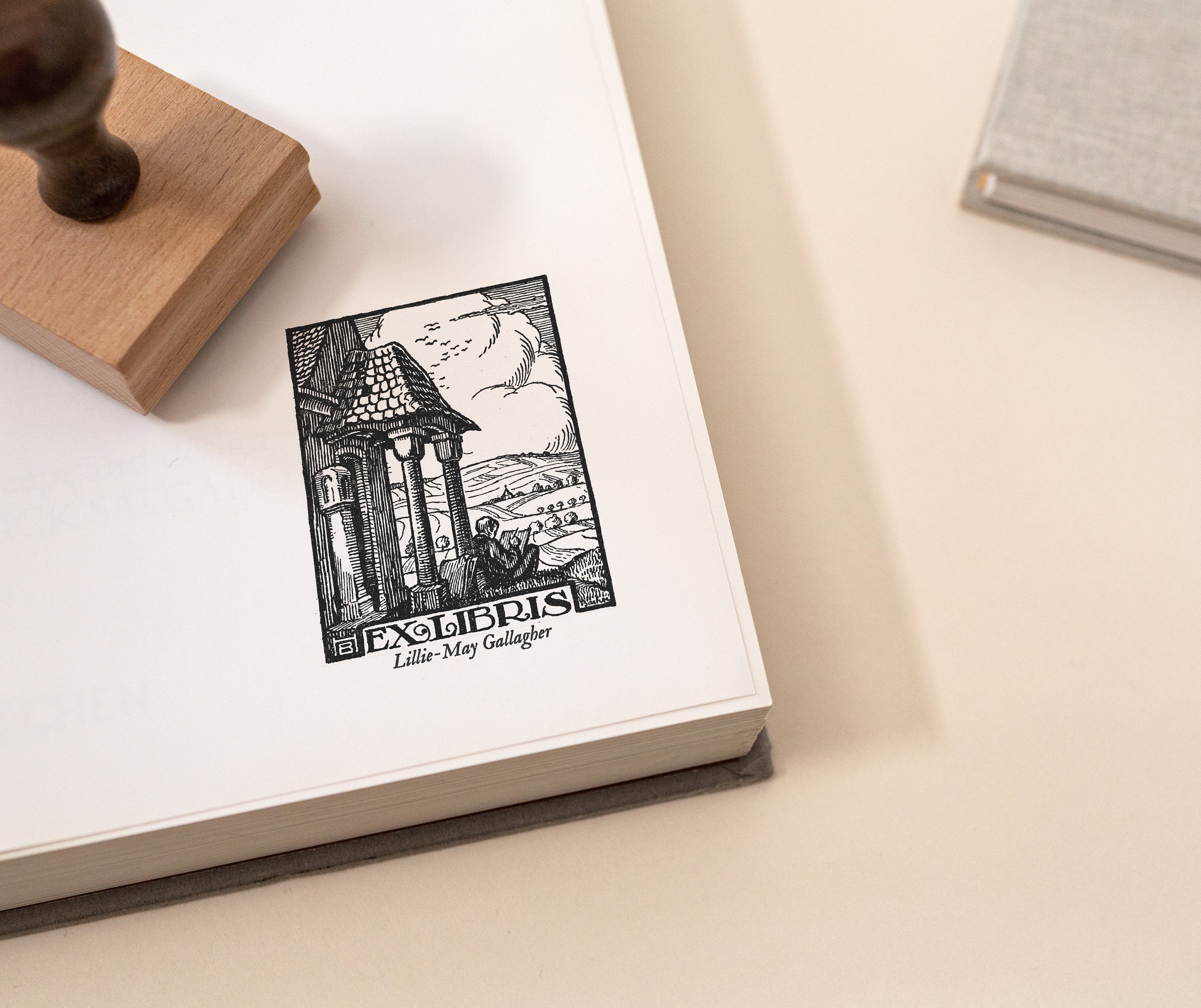 A custom library stamp with a vintage countryside book stamp design, stamped on an open book with a wooden stamp on the side.
