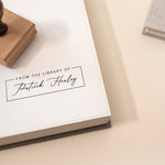 A minimal and modern custom library stamp with the text 'FROM THE LIBRARY OF' and someone's name stamped on white paper in a book.