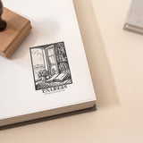 A custom library stamp with a vintage bookplate and a cosy reading nook design is shown being stamped onto an open book with a wooden stamp on the side.
