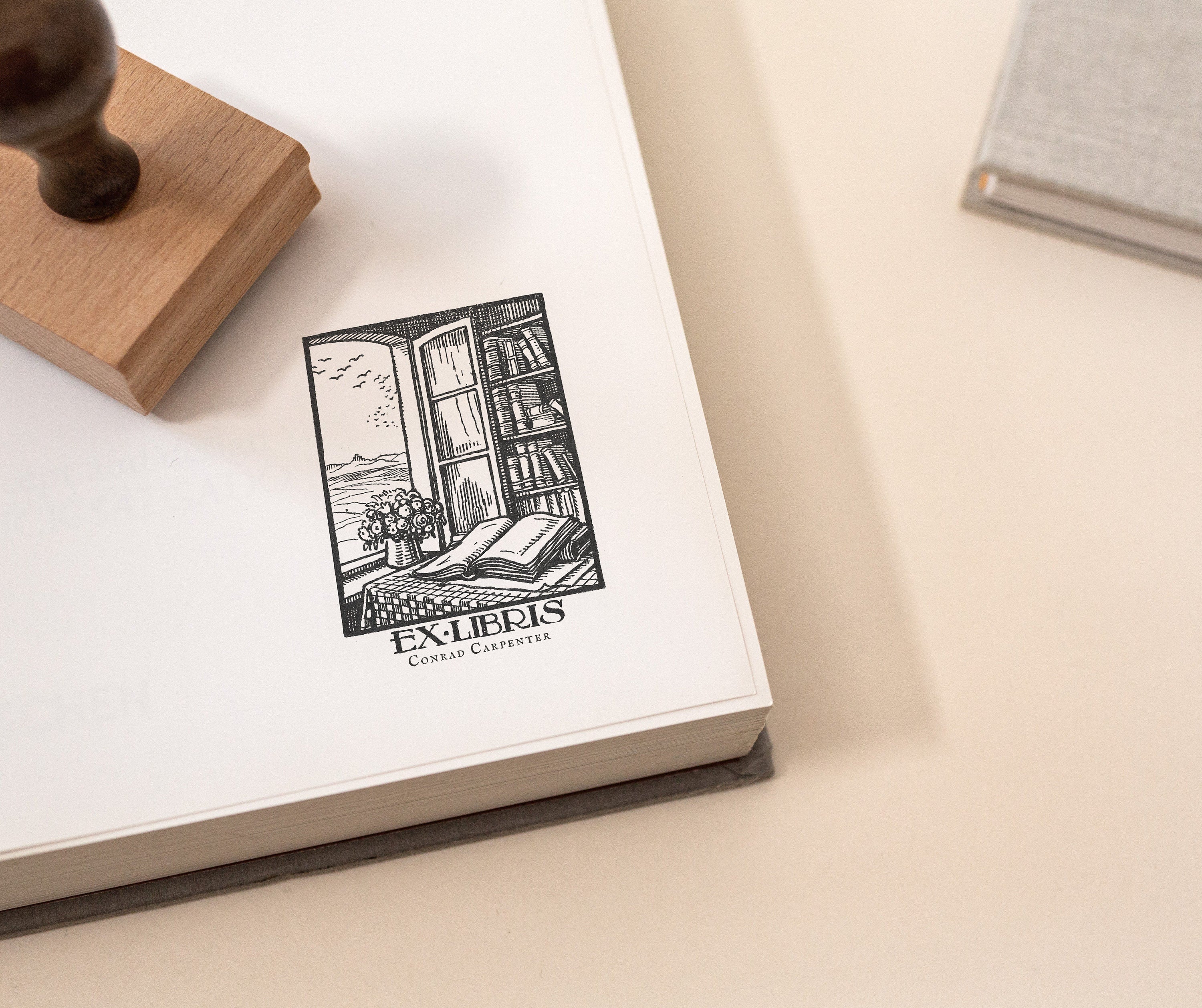 A custom library stamp with a vintage bookplate and a cosy reading nook design is shown being stamped onto an open book with a wooden stamp on the side.
