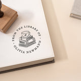 A custom library stamp with a wooden handle, featuring a stack of books illustration and the phrase “From the Library of” followed by your name.