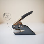 A custom wedding embosser with a black handle and metal body, displayed on a white surface, with a softly blurred background showing the couple’s names, wedding date and a delicate botanical illustration in a circular design.