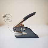 A custom wedding embosser with a black handle and metal body, displayed on a white surface, with a softly blurred background showing the couple’s names, wedding date and a delicate botanical illustration in a circular design.