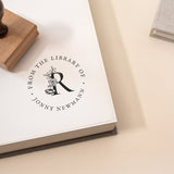 A wooden book stamp with a wooden handle is stamping the phrase 'FROM THE LIBRARY OF' followed by a last name and a floral initial in the middle onto a book page.