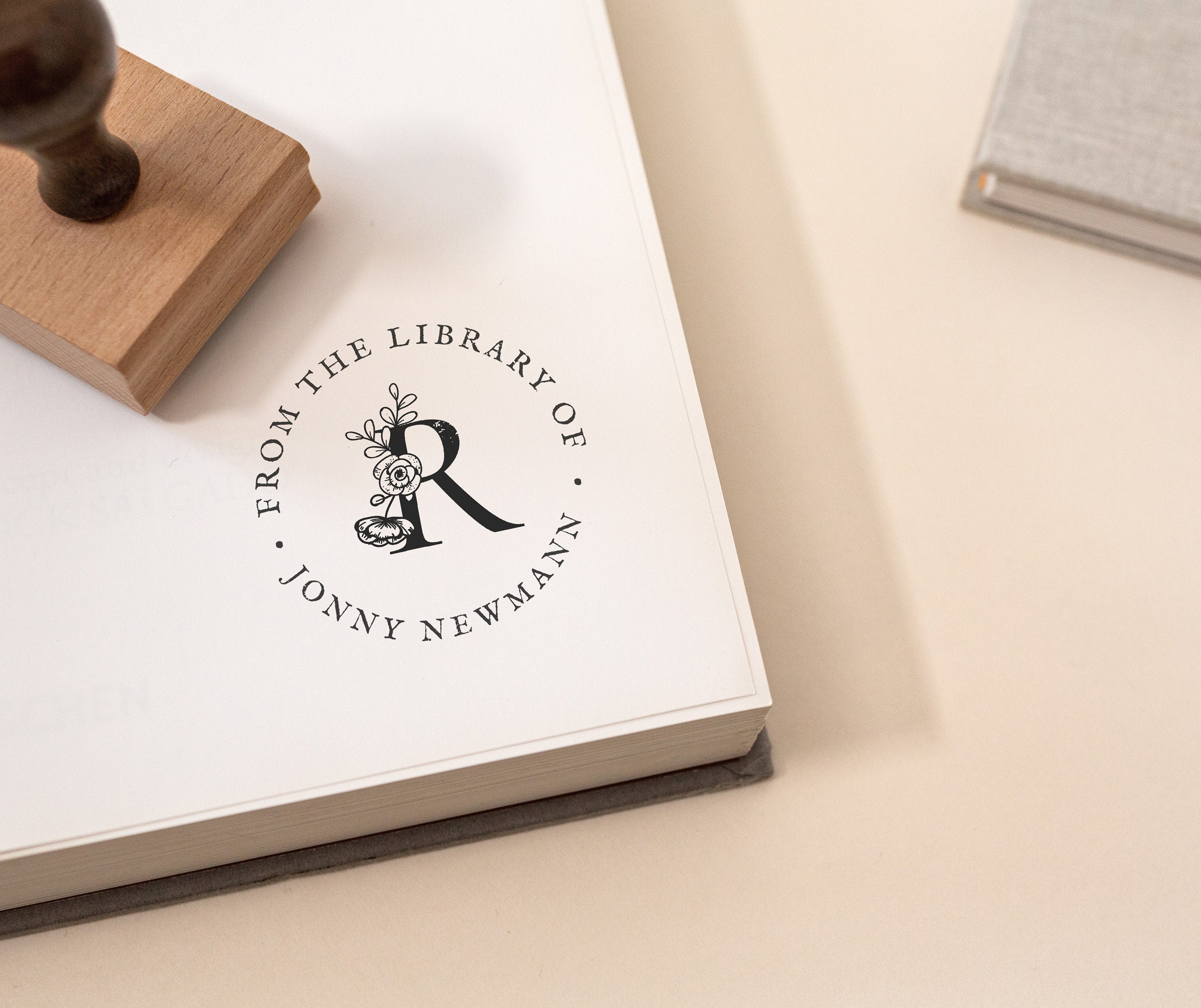 A wooden book stamp with a wooden handle is stamping the phrase 'FROM THE LIBRARY OF' followed by a last name and a floral initial in the middle onto a book page.