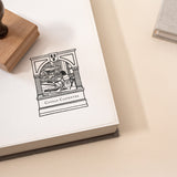 An image of a custom book stamp with a wooden handle resting on an open book, showing a vintage illustration of a library scene.