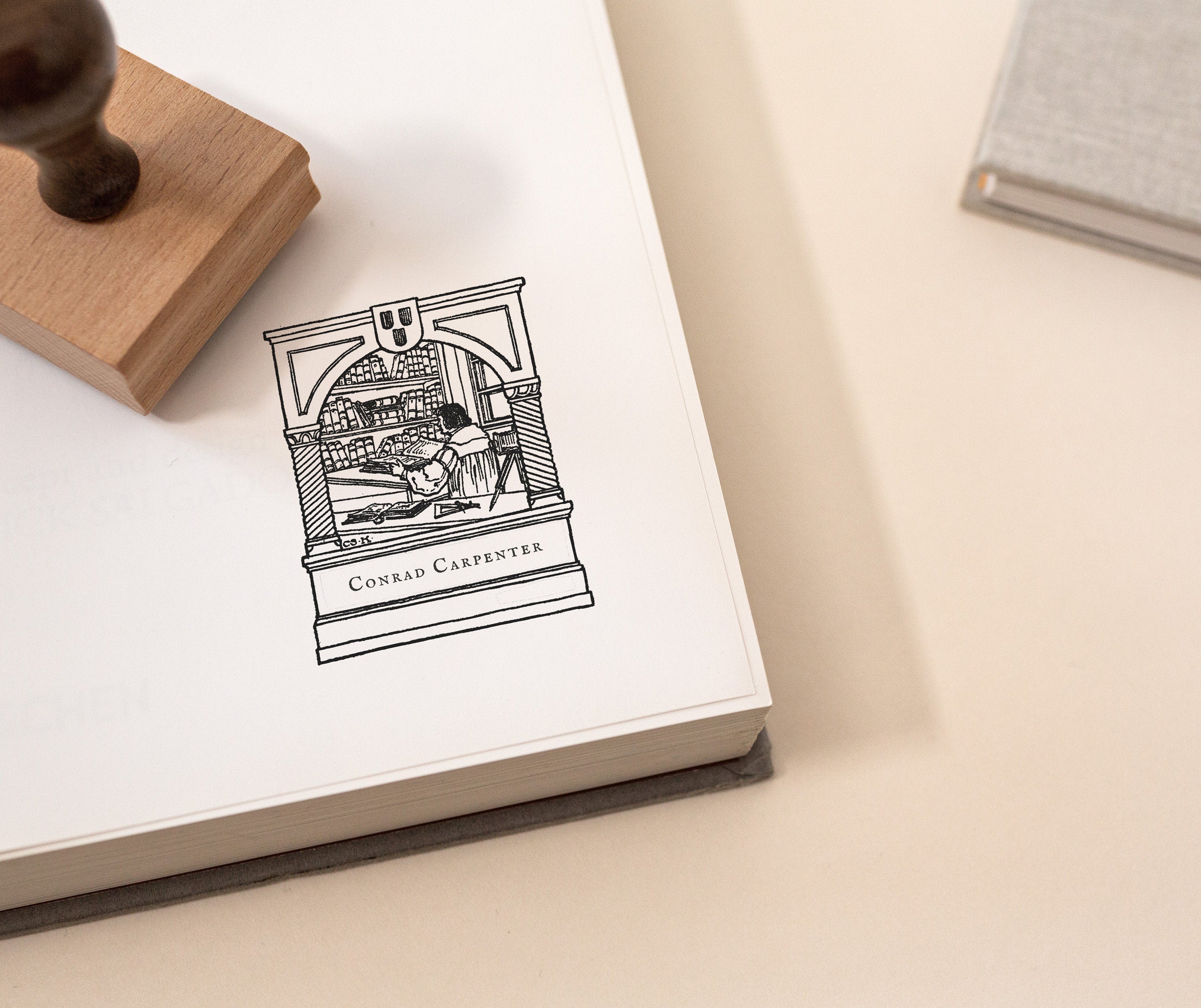 An image of a custom book stamp with a wooden handle resting on an open book, showing a vintage illustration of a library scene.